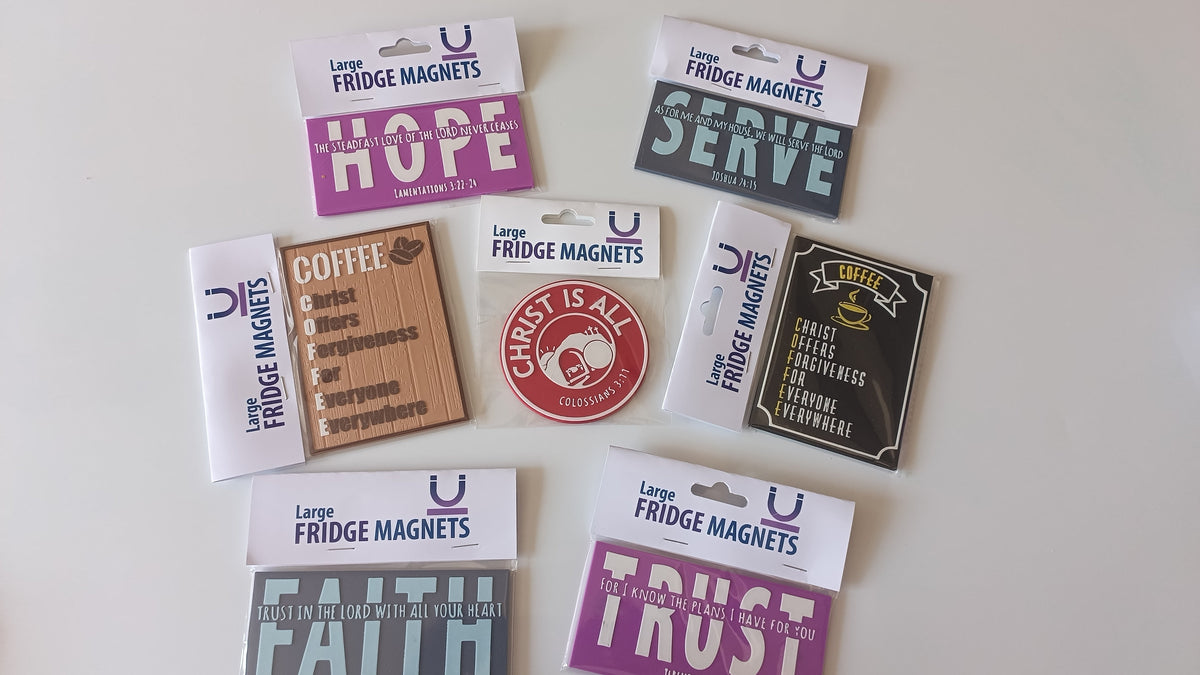 A collection of colorful fridge magnets with religious and inspirational quotes, displayed on a light off-white background. The magnets feature various colors including purple, gray, black, red, brown, and pink, with some still in their white retail packaging.