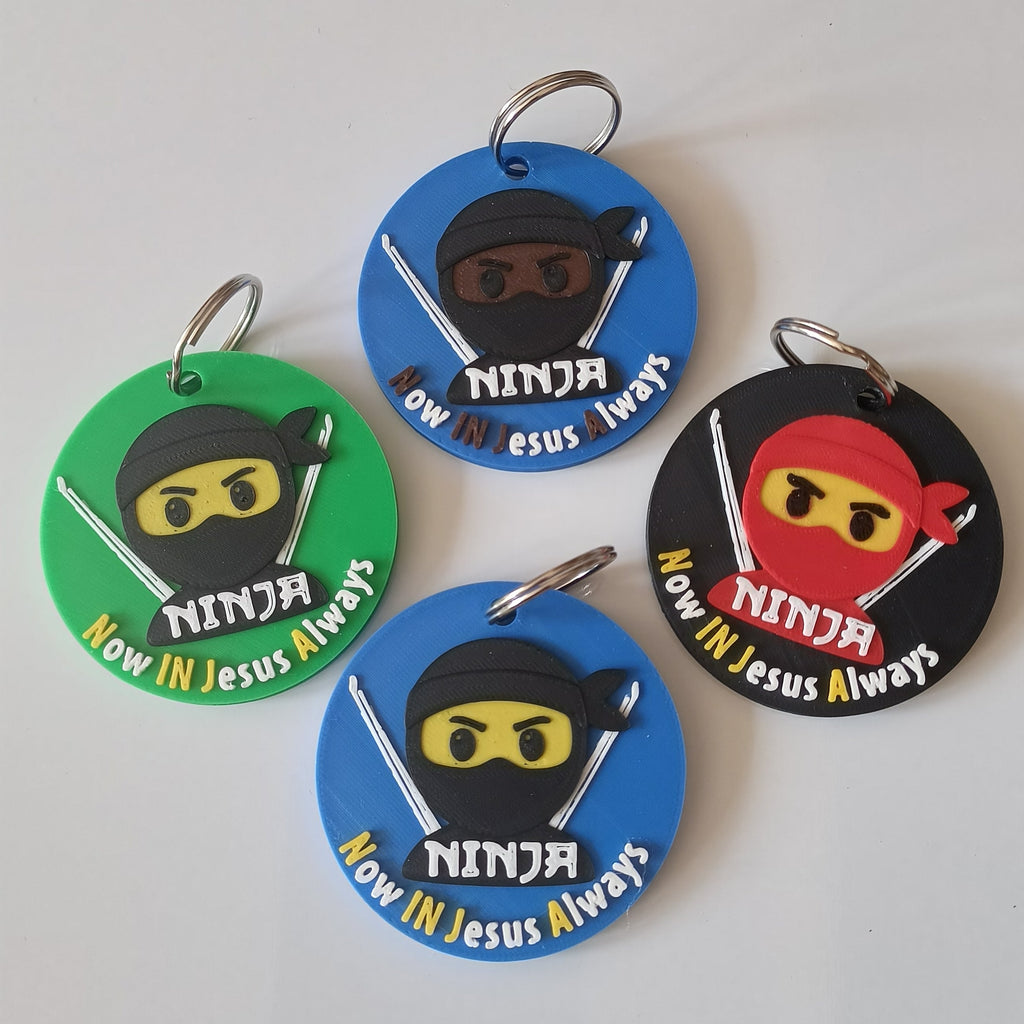 Four circular plastic keychains with metal split rings, featuring cartoon ninja designs and the text 'Now In Jesus Always' on green, blue, black, and blue backgrounds.