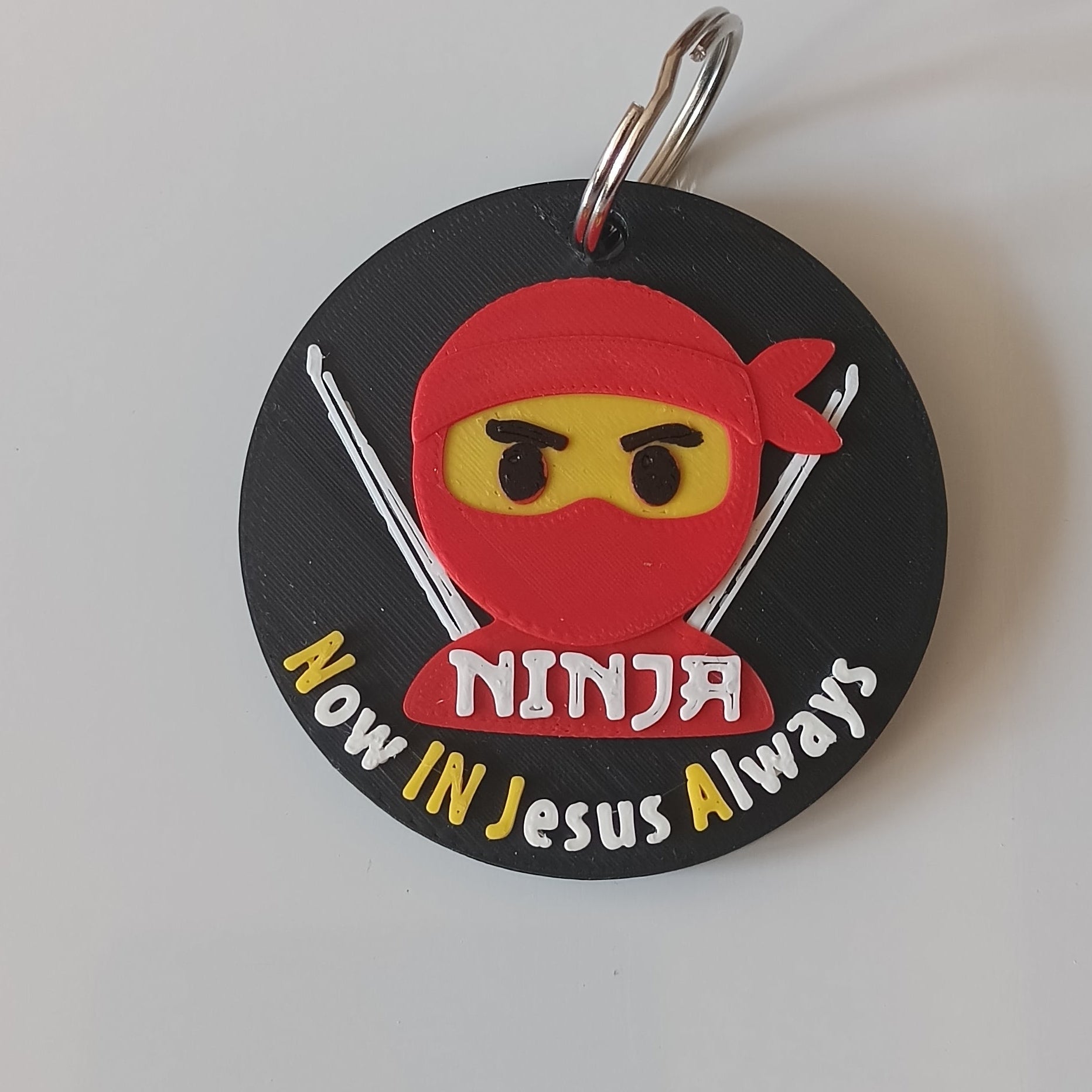 A circular black keychain featuring a cartoon ninja character with a red headband and yellow eyes, with the text 'Now In jesus Always' in white and yellow lettering, attached to a silver metal ring on a plain white background.