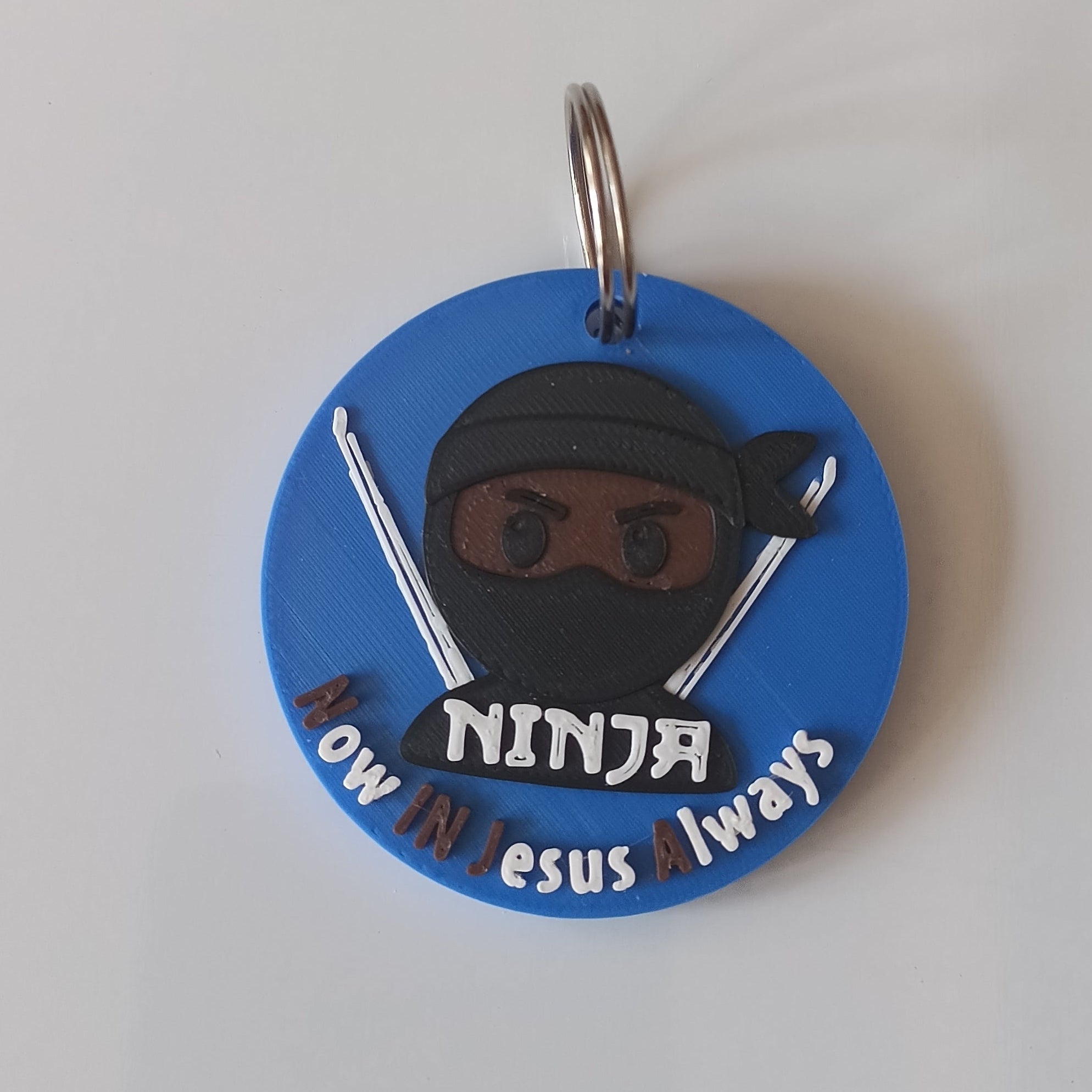 A round blue keychain featuring a cartoon ninja character with the text ''Now In jesus Always'' on a plain white background.