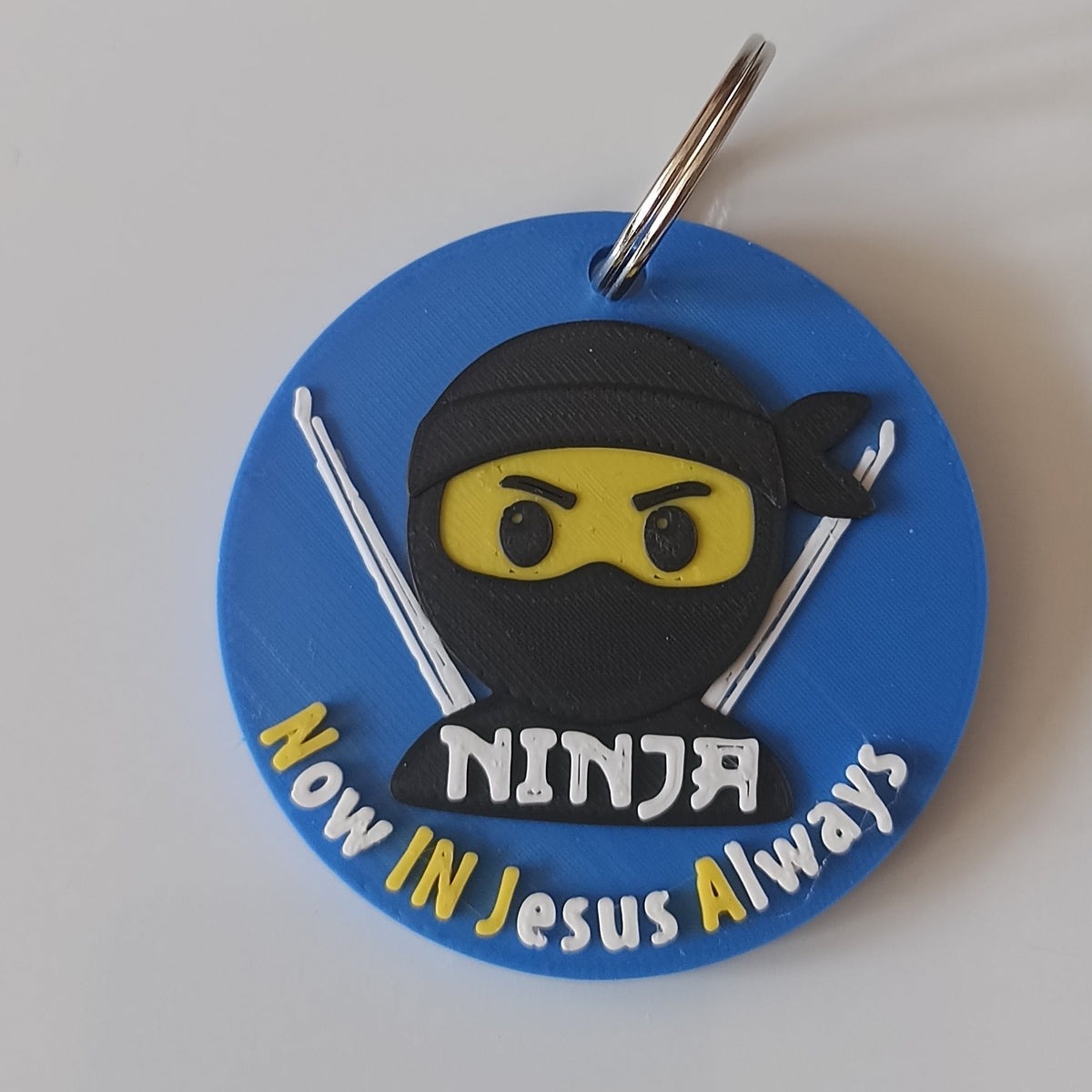 A circular blue keychain featuring a cartoon ninja character with a yellow mask and the text ''Now In jesus Always'' on a plain blue background.