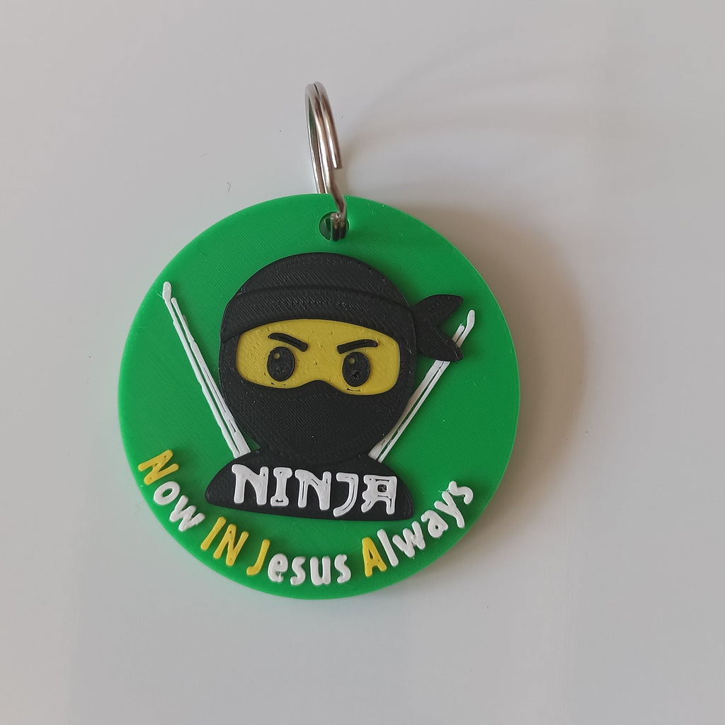 A circular green keychain featuring a cartoon ninja character with a black mask, yellow eyes, and silver swords, with the text ''Now In jesus Always'' in white. The keychain has a metal split ring and is photographed on a plain white surface.
