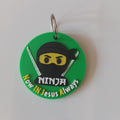 A circular green keychain featuring a cartoon ninja character with a black mask, yellow eyes, and silver swords, with the text ''Now In jesus Always'' in white. The keychain has a metal split ring and is photographed on a plain white surface.