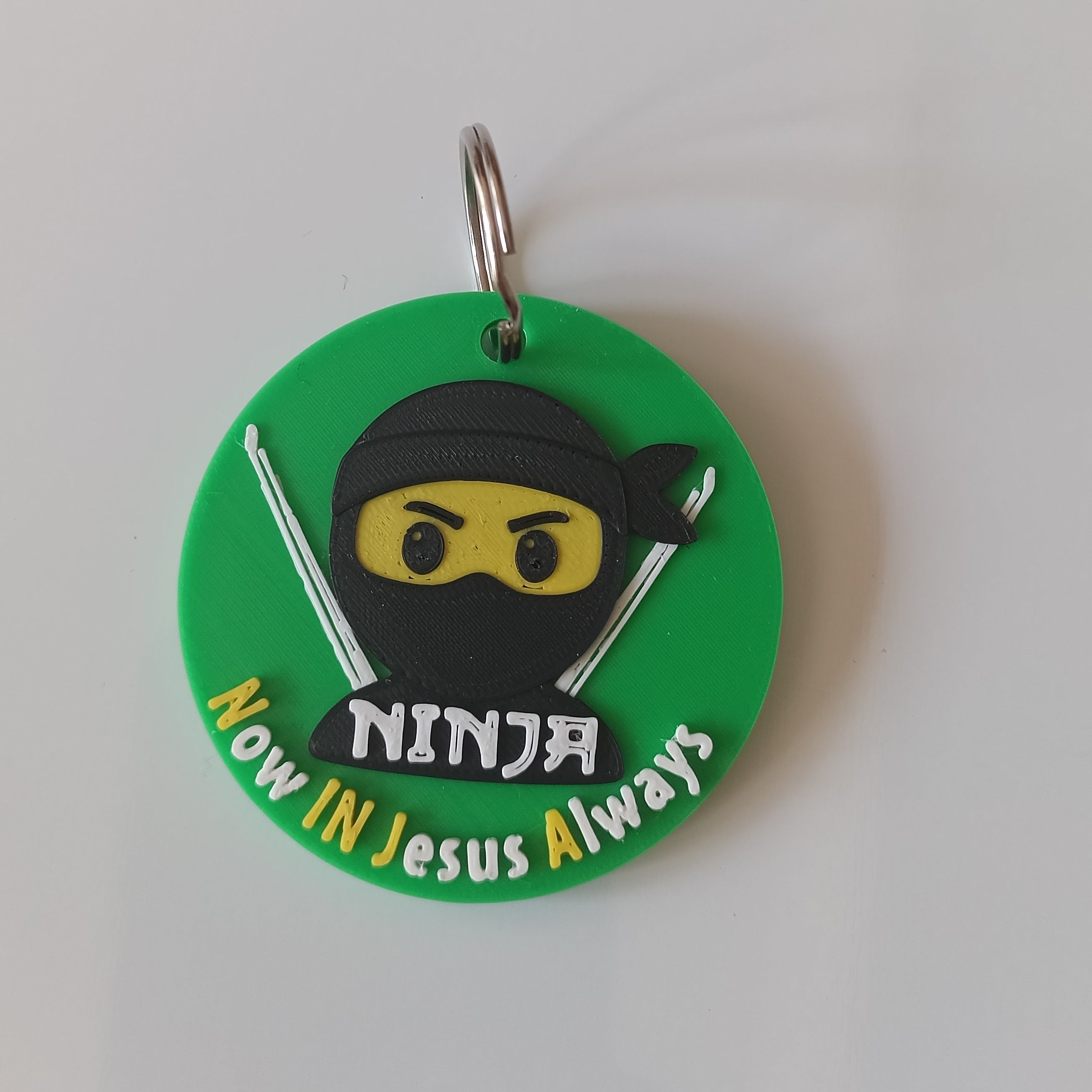 A circular green keychain featuring a cartoon ninja character with a black mask, yellow eyes, and silver swords, with the text ''Now In jesus Always'' in white. The keychain has a metal split ring and is photographed on a plain white surface.