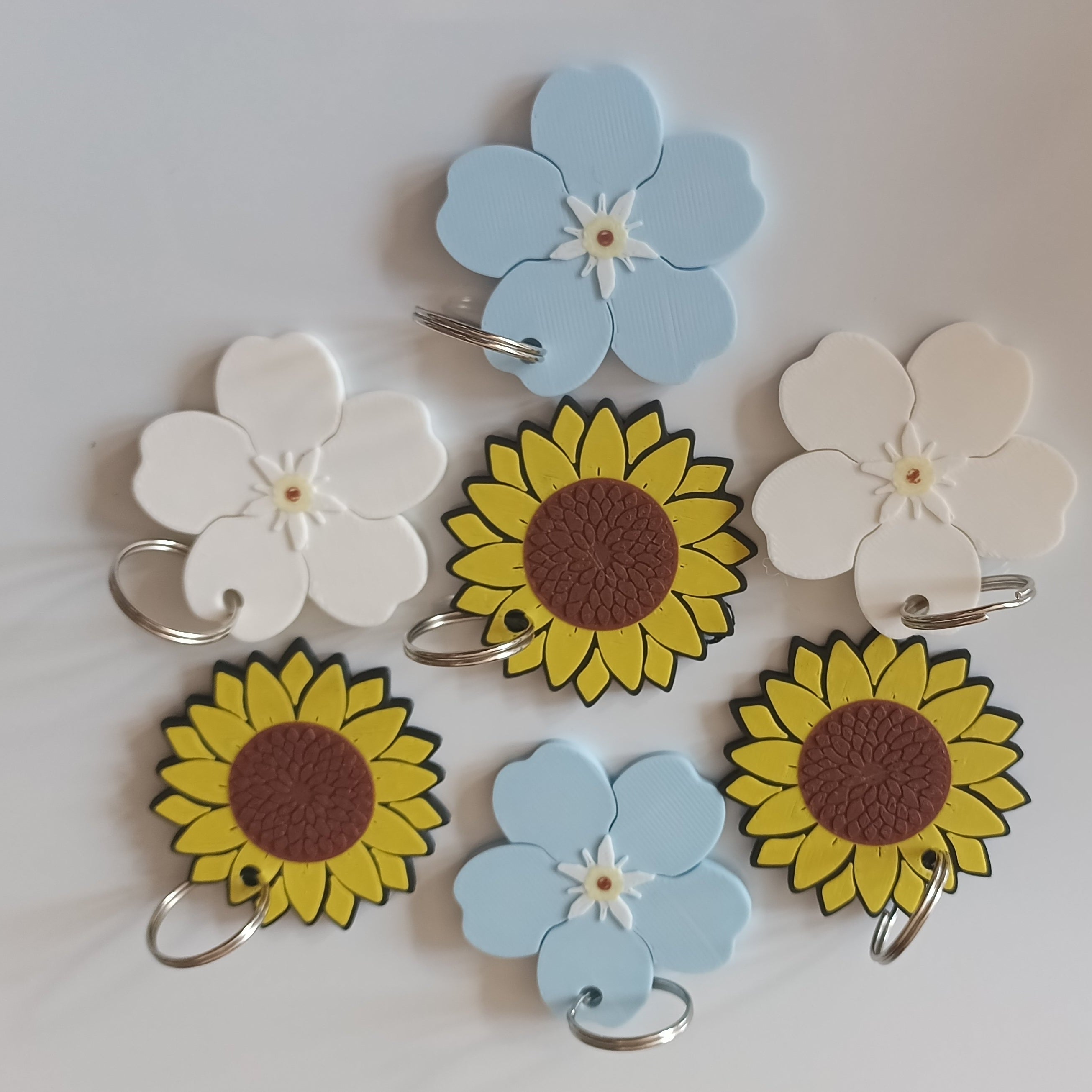 Six decorative flower-shaped clips or tags arranged on a plain white background. The set includes two light blue flowers, two white flowers, and two yellow sunflower designs with brown centers and black outlines, each attached to a small metal ring.