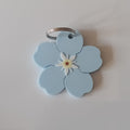 A light blue Forget Me Not flower keychain with a small white and yellow center and a silver metal key ring, shown on a plain light gray background.