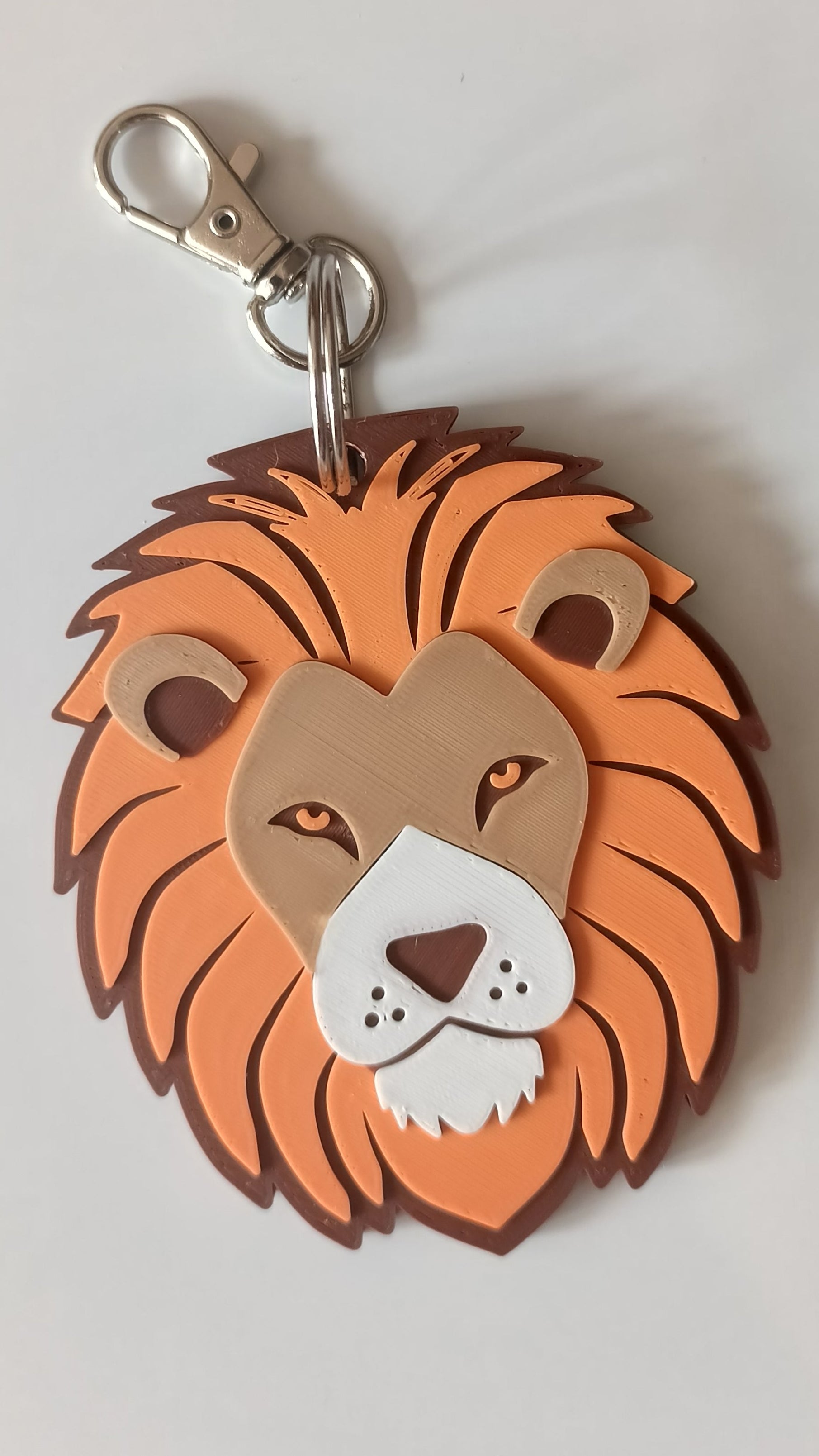 A lion head keychain charm with a metallic silver lobster clasp, featuring layered orange, brown, beige, and white details on a light gray background.