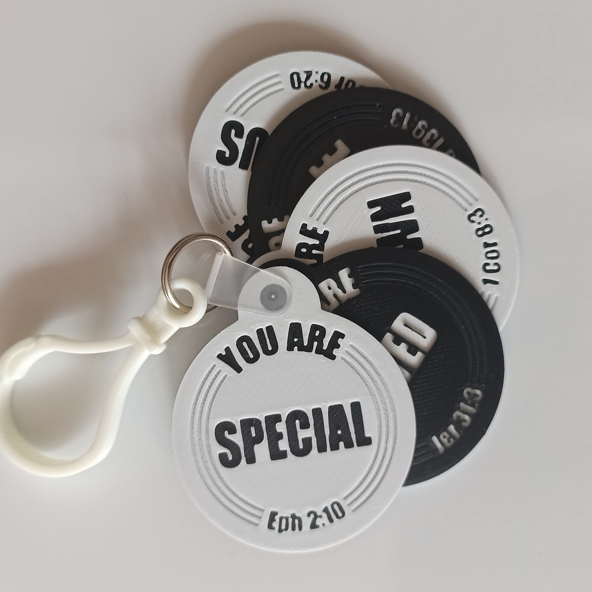 A white plastic keychain with three circular tag charms in white and black, printed with religious and motivational text like 'SPECIAL', 'YOU ARE', and 'EDWN'.
